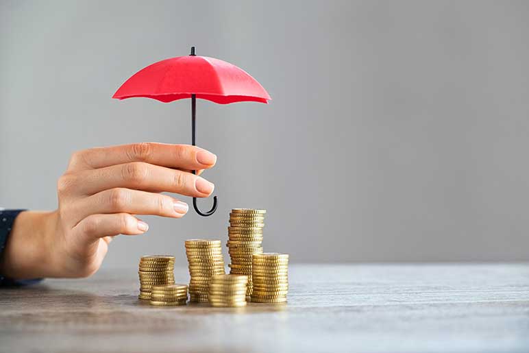 Photograph of human hand holding small red umbrella over several stacks of coins sitting on a table.