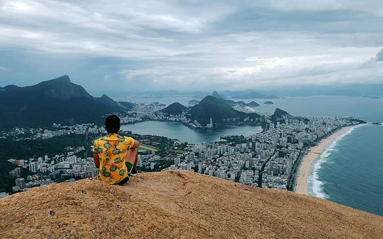 Photograph of a man with back to camera sitting on a high mountain top overlooking a city in Brazil.